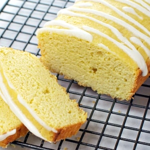 May include: A sliced loaf of yellow cake with white icing drizzled across the top. The cake sits on a black wire cooling rack. One slice is cut and placed in front of the loaf.
