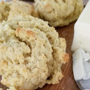 May include: Close-up of freshly baked biscuits with a golden-brown crust. The biscuits are light in color and have a textured surface. A large block of white butter and a butter knife are also visible.