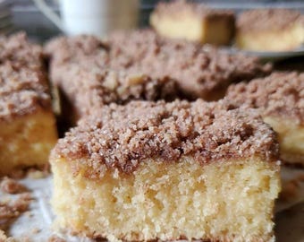 May include: Several slices of coffee cake with a crumb topping are shown. The cake has a light yellow interior and a brown, textured topping. A white mug is visible in the background, suggesting a breakfast or snack setting.
