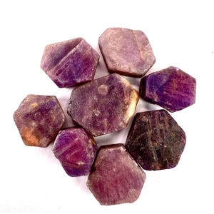 May include: A collection of raw, hexagonal-shaped ruby gemstones. The stones display a range of deep red and purple hues, with some showing lighter, translucent areas. The gemstones are arranged on a white surface.