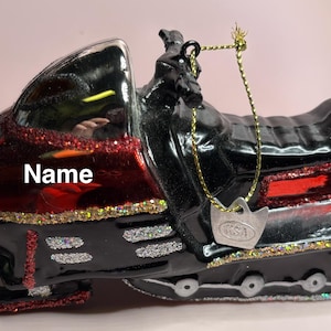 Puede incluir: Adorno decorativo de motonieve en negro y rojo, con detalles brillantes y una cuerda dorada para colgar. La palabra "Name" es visible en el lateral.