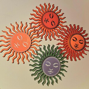 Puede incluir: Cuatro ilustraciones estilizadas de soles con ojos cerrados y varios colores. Los soles tienen caras naranjas, rojas, verdes y moradas con rayos ondulados. El fondo es de color beige claro.