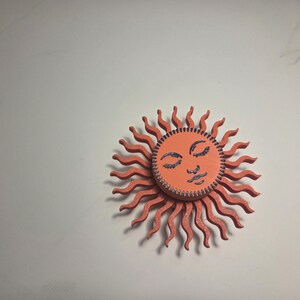 Puede incluir: Adorno de pared de madera con forma de sol sonriente. El sol tiene un cuerpo de color coral con rayos ondulados y una cara naranja. La cara presenta ojos cerrados y un dise&ntilde;o cosido.