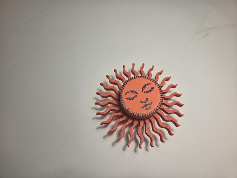 Puede incluir: Adorno de pared de madera con forma de sol sonriente. El sol tiene un cuerpo de color coral con rayos ondulados y una cara naranja. La cara presenta ojos cerrados y un dise&ntilde;o cosido.