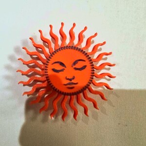 Puede incluir: Un objeto decorativo en forma de sol naranja con una cara y rayos ondulados. El sol tiene los ojos cerrados, una nariz peque&ntilde;a y una boca sonriente. La cara del sol es de un naranja m&aacute;s oscuro que los rayos.