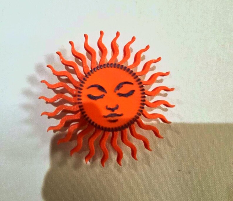 Puede incluir: Un objeto decorativo en forma de sol naranja con una cara y rayos ondulados. El sol tiene los ojos cerrados, una nariz peque&ntilde;a y una boca sonriente. La cara del sol es de un naranja m&aacute;s oscuro que los rayos.