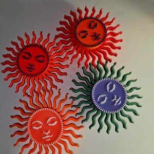 Puede incluir: Cuatro adornos de pared con forma de sol. Cada sol tiene una cara circular con los ojos cerrados y una boca sonriente. Los soles son en tonos de rojo, naranja, verde y morado. Los rayos de los soles son ondulados y se extienden hacia afuera de la cara.