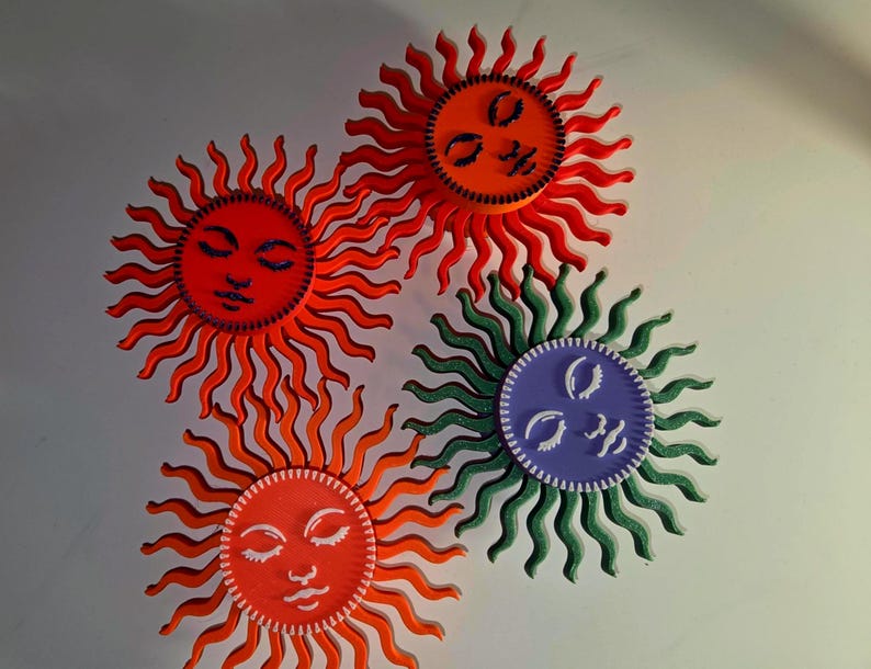 Puede incluir: Cuatro adornos de pared con forma de sol. Cada sol tiene una cara circular con los ojos cerrados y una boca sonriente. Los soles son en tonos de rojo, naranja, verde y morado. Los rayos de los soles son ondulados y se extienden hacia afuera de la cara.
