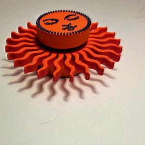 Puede incluir: Objeto decorativo en forma de sol naranja con una cara sonriente en la parte superior. El sol tiene rayos ondulados y un contorno negro. La cara es negra y tiene los ojos cerrados y una boca sonriente. El objeto est&aacute; sobre una superficie blanca.