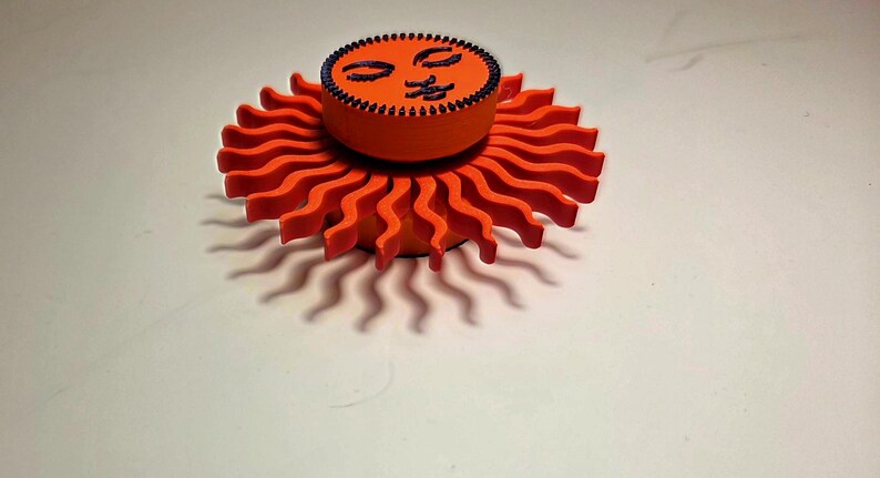 Puede incluir: Objeto decorativo en forma de sol naranja con una cara sonriente en la parte superior. El sol tiene rayos ondulados y un contorno negro. La cara es negra y tiene los ojos cerrados y una boca sonriente. El objeto est&aacute; sobre una superficie blanca.