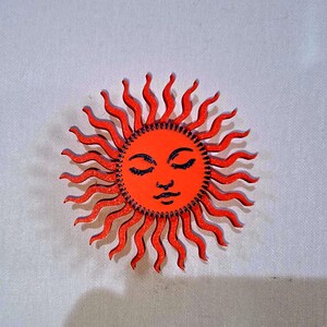 Puede incluir: Un dise&ntilde;o de sol de madera con una cara sonriente y ojos cerrados. El sol est&aacute; pintado de naranja con rayos ondulados y detalles negros. El sol mide aproximadamente 10 cm de di&aacute;metro.