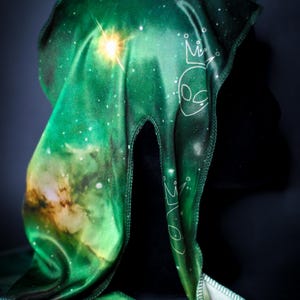 May include: Green durag with a galaxy print, featuring white alien head and crown graphics. The fabric has a silky sheen and a vibrant color palette of greens, blues, and yellows. The durag is designed to be worn on the head.