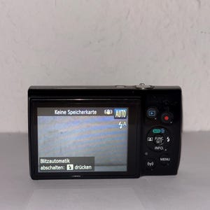 May include: A black digital camera with a screen displaying German text. The screen shows "Keine Speicherkarte" and "Blitzautomatik abschalten: drücken". The camera has several buttons, including "AUTO", "FUNC. SET", and "MENU".