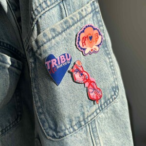 Può includere: Primo piano di una giacca di jeans azzurra con tre spille colorate. Le spille presentano le parole "AMOUR" e "TRIBU sororie" in rosa e blu, con disegni floreali e a forma di cuore.