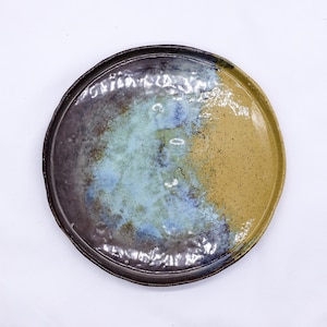 May include: A handcrafted ceramic plate with a distinctive colour palette. It has a dark brown rim, blending into a section of muted blue and a speckled tan area. The plate has an organic, irregular shape, with a rustic feel.