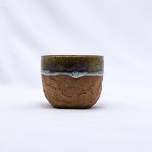 K&ouml;nnte beinhalten: Eine kleine, handgefertigte Keramiktasse mit einer strukturierten, braunen Au&szlig;enseite. Der Rand ist dunkel olivgr&uuml;n mit einer wei&szlig;en und blauen Glasur. Die Tasse wird wahrscheinlich zum Trinken von Getr&auml;nken verwendet.