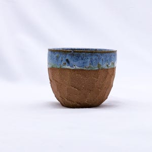 K&ouml;nnte beinhalten: Eine handgefertigte Keramiktasse mit einem strukturierten, braunen Boden und einem blau glasierten Rand. Die Tasse hat ein rustikales, organisches Aussehen, perfekt f&uuml;r Kaffee oder Tee. Die blaue Glasur hat einen gesprenkelten Effekt.