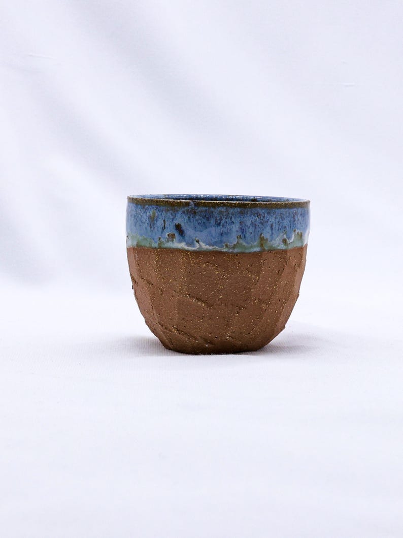 K&ouml;nnte beinhalten: Eine handgefertigte Keramiktasse mit einem strukturierten, braunen Boden und einem blau glasierten Rand. Die Tasse hat ein rustikales, organisches Aussehen, perfekt f&uuml;r Kaffee oder Tee. Die blaue Glasur hat einen gesprenkelten Effekt.