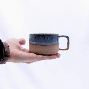 May include: A handcrafted ceramic mug with a two-tone design. The top half is a deep blue glaze, the bottom half is a natural brown. The mug has a square handle. It is held in a hand against a white background.