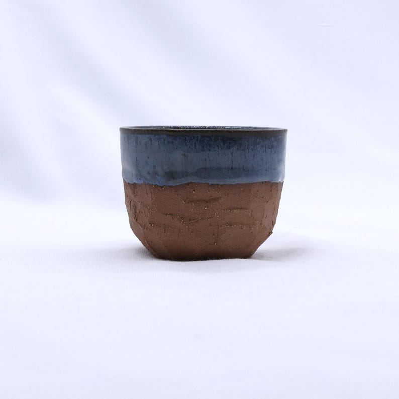 K&ouml;nnte beinhalten: Eine handgefertigte Keramikschale mit zweifarbigem Design. Die obere H&auml;lfte ist in einem tiefen Blau glasiert, w&auml;hrend die untere H&auml;lfte eine strukturierte, erdfarbene braune Oberfl&auml;che aufweist. Die Schale hat eine abgerundete Form und eine rustikale &Auml;sthetik.