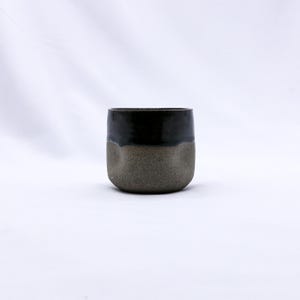 K&ouml;nnte beinhalten: Handgefertigte Keramiktasse mit zweifarbigem Design. Die obere H&auml;lfte ist gl&auml;nzend schwarz, kontrastierend mit der strukturierten, erdig-graubraunen unteren H&auml;lfte. Die Tasse hat eine abgerundete, minimalistische Form, geeignet f&uuml;r Getr&auml;nke.