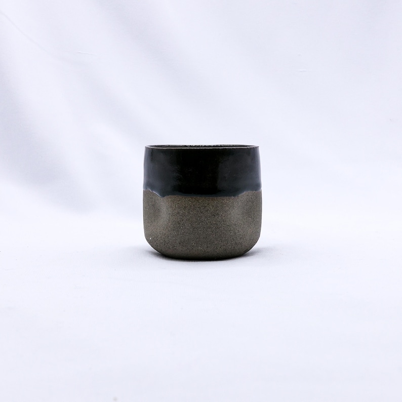 K&ouml;nnte beinhalten: Handgefertigte Keramiktasse mit zweifarbigem Design. Die obere H&auml;lfte ist gl&auml;nzend schwarz, kontrastierend mit der strukturierten, erdig-graubraunen unteren H&auml;lfte. Die Tasse hat eine abgerundete, minimalistische Form, geeignet f&uuml;r Getr&auml;nke.