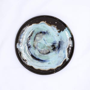 May include: A round ceramic plate with a dark brown rim and a light blue glaze with a swirling pattern in the centre.
