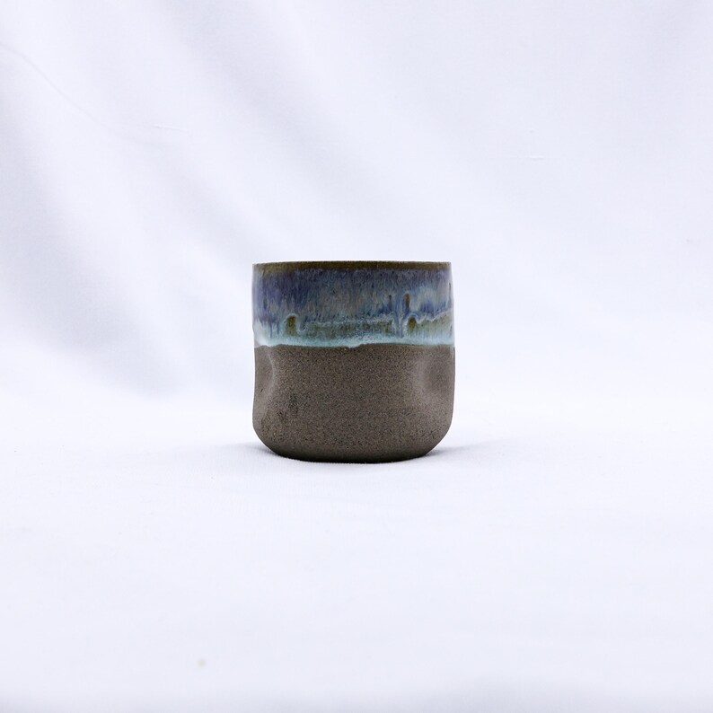 K&ouml;nnte beinhalten: Eine handgefertigte Keramiktasse mit einem einzigartigen, strukturierten Design. Die Tasse hat einen dunkelbraunen Boden und einen oberen Bereich mit einer blauen und gr&uuml;nen Glasur. Die Glasur hat einen Tropfeffekt, der den handwerklichen Reiz der Tasse unterstreicht.