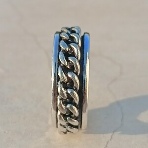 May include: A silver ring featuring a chain design. The ring is made of two bands, one with a smooth surface and the other with a chain design.