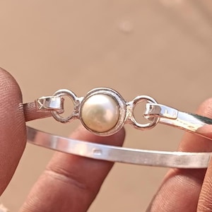 Op de afbeelding: Een zilveren armband met een enkele witte parel in het midden. De armband heeft een eenvoudig ontwerp met twee kleine lussen aan weerszijden van de parel.