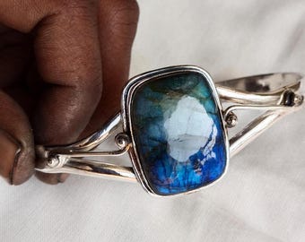 Puede incluir: Una pulsera de plata con una piedra preciosa grande, rectangular, azul y verde. La piedra preciosa tiene un efecto brillante e iridiscente.