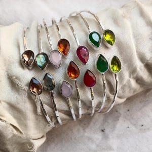 Può includere: Un set di braccialetti d'argento con accenti di pietre preziose colorate. Ogni braccialetto presenta una pietra preziosa a forma di lacrima di un colore diverso, tra cui rosa, rosso, verde, marrone e trasparente.