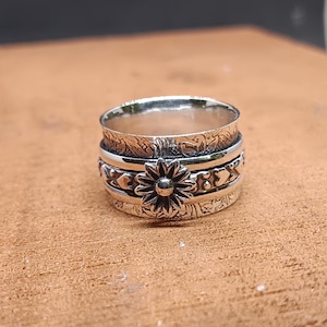 May include: A silver spinning ring featuring a flower design and other decorative elements. The ring is made of multiple bands that spin freely.