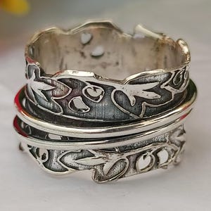 Könnte beinhalten: Ein silberner Ring mit einem floralen Design. Der Ring besteht aus zwei Bändern, die übereinander liegen. Das obere Band hat ein filigraneres Design mit Blättern und Blumen. Das untere Band ist ein einfaches Band.