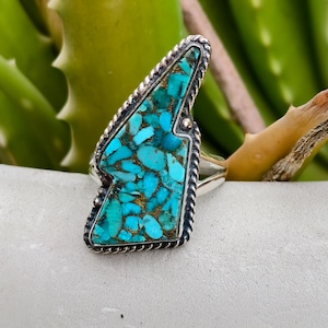 Op de afbeelding: Een zilveren ring met een grote, turquoise gekleurde steen. De steen is een mozaïek van blauwe en gouden tinten, gezet in een zilveren frame met een touwachtige rand. De ring heeft een unieke, hoekige vorm.