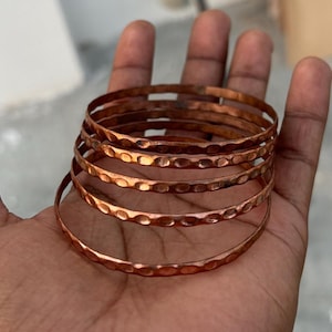 Può includere: Un set di cinque braccialetti in rame con una texture martellata. I braccialetti sono impilati uno sopra l'altro e sono di un colore marrone rossiccio caldo.