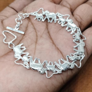 Könnte beinhalten: Ein silbernes Armband mit einer Kette und einer Reihe von Elefantenanhängern. Die Elefanten sind alle in die gleiche Richtung ausgerichtet und durch ihre Rüssel mit der Kette verbunden.
