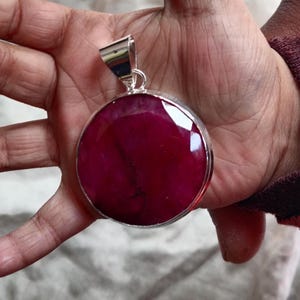 Op de afbeelding: Een ronde, dieprode edelsteen hanger met een zilveren zetting. De hanger wordt in een hand gehouden.