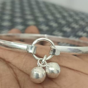 May include: A silver bangle bracelet with a round charm attached. The charm has two small bells hanging from it.