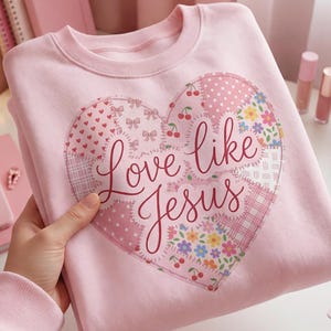 Puede incluir: Una sudadera rosa claro con un diseño en forma de corazón. El corazón está compuesto por patrones de patchwork con cerezas, lazos y detalles florales. Las palabras "Love like Jesus" están escritas en cursiva sobre el corazón.
