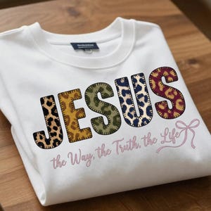 Puede incluir: Sudadera blanca con la palabra "JESUS" en un diseño de patchwork con estampados de leopardo en varios colores. Debajo de la palabra "JESUS" está el texto "the Way, the Truth, the Life" en una fuente rosa claro.