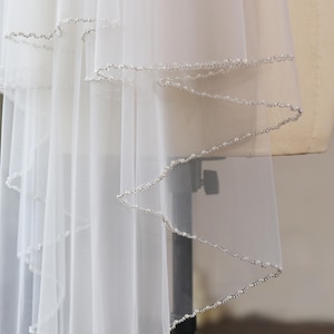 Op de afbeelding: Een meerlaagse witte tule sluier met een geschulpte rand, afgewerkt met delicate zilveren kraaltjes en kleine witte parels. De transparante stof valt in zachte plooien en toont de gedetailleerde kralenafwerking langs de gebogen zoom.