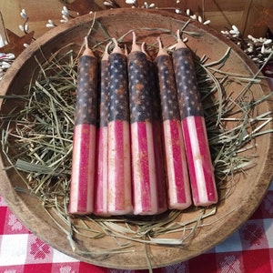 May include: A set of six red, white, and blue candles with an American flag design. The candles are in a wooden bowl filled with hay.