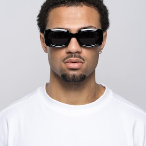 May include: Black rectangular sunglasses with dark lenses. The sunglasses have a thick frame and are worn by a person wearing a white crew neck t-shirt. The background is a solid light grey.