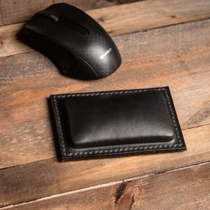 May include: A black leather mouse pad with a stitched border and a raised center. The mouse pad is on a wooden surface.