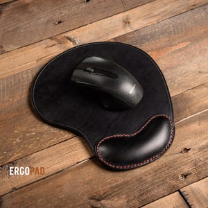 Può includere: Tappetino per mouse in pelle nera con un poggiapolsi e un mouse nero in cima. Il tappetino per mouse ha cuciture rosse attorno ai bordi. Il testo "ERCOPAD" è visibile nell'angolo in basso a sinistra dell'immagine.
