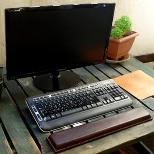 Könnte beinhalten: Ein schwarzer Computermonitor mit Samsung-Logo steht auf einem Holztisch. Eine schwarze Microsoft-Tastatur und eine braune Leder-Handgelenkauflage befinden sich vor dem Monitor. Eine kleine Topfpflanze ist im Hintergrund zu sehen.