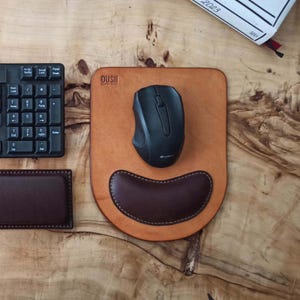 Può includere: Un tappetino per mouse in pelle con un poggiapolsi marrone e un mouse per computer nero. Sul tappetino è stampata la parola "OUSHII". Sono visibili anche una tastiera nera e un piccolo cuscinetto in pelle.