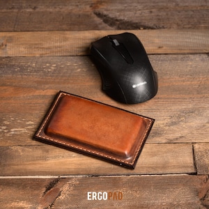 May include: A brown leather mouse pad with a stitched border. The mouse pad is on a wooden surface with a black wireless mouse on top of it. The mouse has the Logitech logo on it.