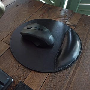 May include: A black leather mouse pad with a built-in wrist rest and a black wireless mouse. The mouse pad is round with a stitched edge. The mouse has a scroll wheel and buttons.